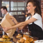 A woman stands behind a bakery counter. She's handing over a paper bag to a customer we can't see. (Fair Work Act)