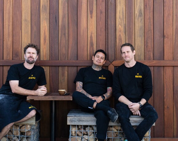 The three The Bread Social owners sit in front of a timber wall. They each wear black shirts and aprons. They are looking at the camera without smiling