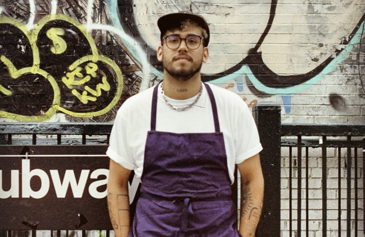 Tony Yamera from Bakery by Textbook stands in front of a graffitied NYC subway sign. He has blond hair, and wears glasses. He has on a white t-shirt with a purple apron over the top.