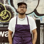 Tony Yamera from Bakery by Textbook stands in front of a graffitied NYC subway sign. He has blond hair, and wears glasses. He has on a white t-shirt with a purple apron over the top.