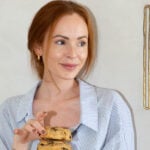 Brooke Saward, a woman with red hair falling loosely around her face, stands in front a white wall. She looks to the right of the camera and is softly smiling