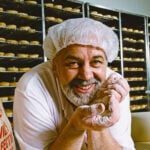 Vilmos ‘Vili’ Milisits from Cafe De Vili's leans over a counter. He wears a hair net and has a grey beard. He is smiling at the camera while holding a handful of flour