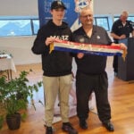 Liam Dyke wears a black hoodie and cream long pants and a black cap. He stands with a man wearing black pants and t-shirt. they are both holding a ribbon from the Tasmanian Baking Show