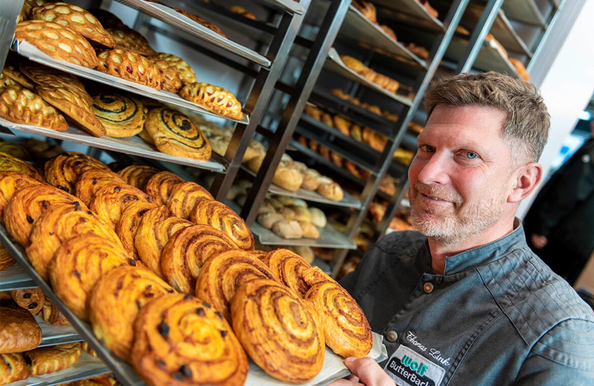 iba will be held again in Dusseldorf next month. Pictured is a man holding a tray of scrolls. He wears a black shirt and is smiling at the camera.