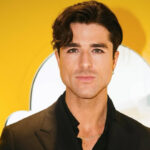 Donut Daddy aka Anthony Randello-Jahn will be attending the Baking Industry Trade Show in June. Pictured is Anthony, a white man with short dark hair and dark eyes. He's wearing a black suit and standing in front of a bright yellow background. He's not smiling.