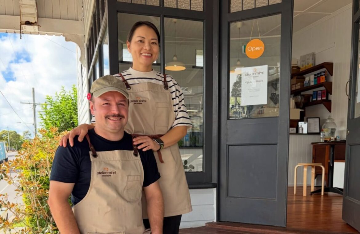 Paul and Miki from newly opened Atelier Mimi. Pictured is a man and woman standing on wooden stairs. The man is white with dark hair. He's earing a tan apron and cap. The woman has dark hair and is wearing a striped shirt and tan apron. They are both smiling at the camera.
