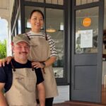 Paul and Miki from newly opened Atelier Mimi. Pictured is a man and woman standing on wooden stairs. The man is white with dark hair. He's earing a tan apron and cap. The woman has dark hair and is wearing a striped shirt and tan apron. They are both smiling at the camera.