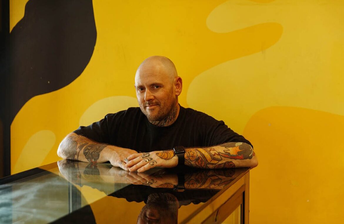 Jason Spencer from Banana Boogie Bakery, which has just been named Australia's Best Bakery. Pictured is a bald, white man wearing a black shirt and with tattoos down his arms. He is standing behind a glass counter and in front of a bright yellow wall. He is looking at the camera.
