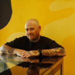 Jason Spencer from Banana Boogie Bakery, which has just been named Australia's Best Bakery. Pictured is a bald, white man wearing a black shirt and with tattoos down his arms. He is standing behind a glass counter and in front of a bright yellow wall. He is looking at the camera.