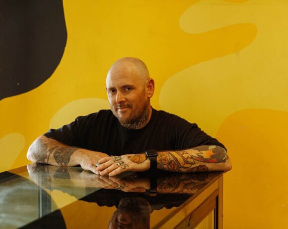 Jason Spencer from Banana Boogie Bakery, which has just been named Australia's Best Bakery. Pictured is a bald, white man wearing a black shirt and with tattoos down his arms. He is standing behind a glass counter and in front of a bright yellow wall. He is looking at the camera.