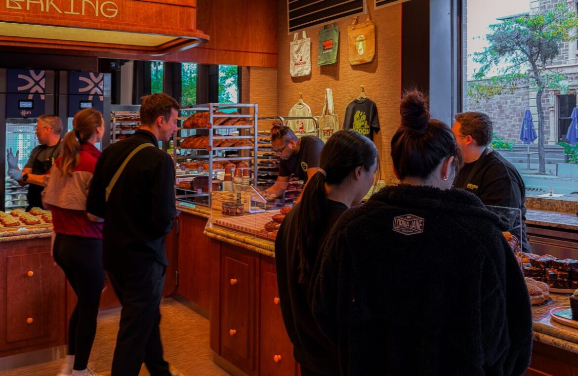 Shadow Baking's newest location in North Adelaide has opened its doors. Pictured is a warm bakery with wooden counters and warm lighting. It is full of people looking at glass counters full of baked goods