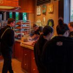 Shadow Baking's newest location in North Adelaide has opened its doors. Pictured is a warm bakery with wooden counters and warm lighting. It is full of people looking at glass counters full of baked goods