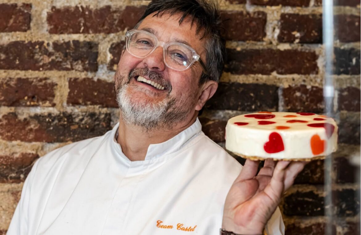Benoît Castel. Pictured is a man in chef whites against a brick wall. He's holding a white cake decorated with strawberries. He has greying hair and glasses and is smiling at the camera