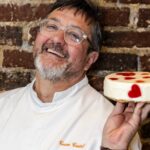 Benoît Castel. Pictured is a man in chef whites against a brick wall. He's holding a white cake decorated with strawberries. He has greying hair and glasses and is smiling at the camera